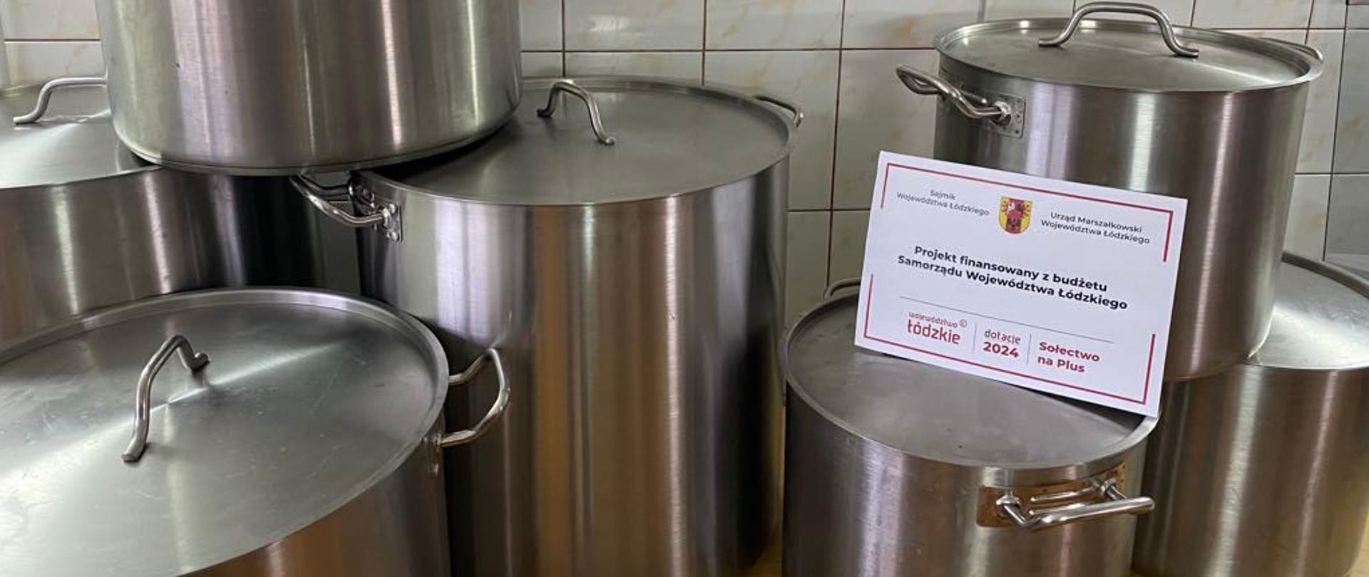 Na zdjęciu znajduje się kilka dużych, metalowych garnków ustawionych na blacie w kuchni. Garnki mają błyszczące, stalowe powierzchnie i różnią się rozmiarami. Wśród nich leży również karta informacyjna, na której widnieje napis dotyczący projektu finansowanego z budżetu Samorządu Województwa Łódzkiego. Tło zdjęcia stanowią jasne płytki ceramiczne.