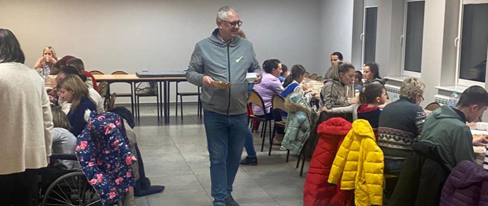 ludzie siedzący na krzesłach przy stołach w sali Miejsko-Gminnego Ośrodka Kultury w Margoninie, dwie osoby stoją