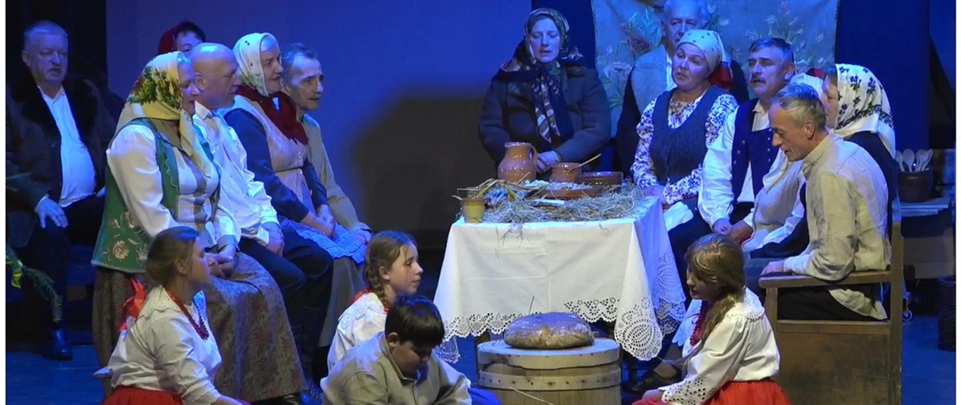 kobiety, mężczyźni i dzieci w starych ludowych strojach siedzą przy wspólnym stole, dzieci na podłodze 