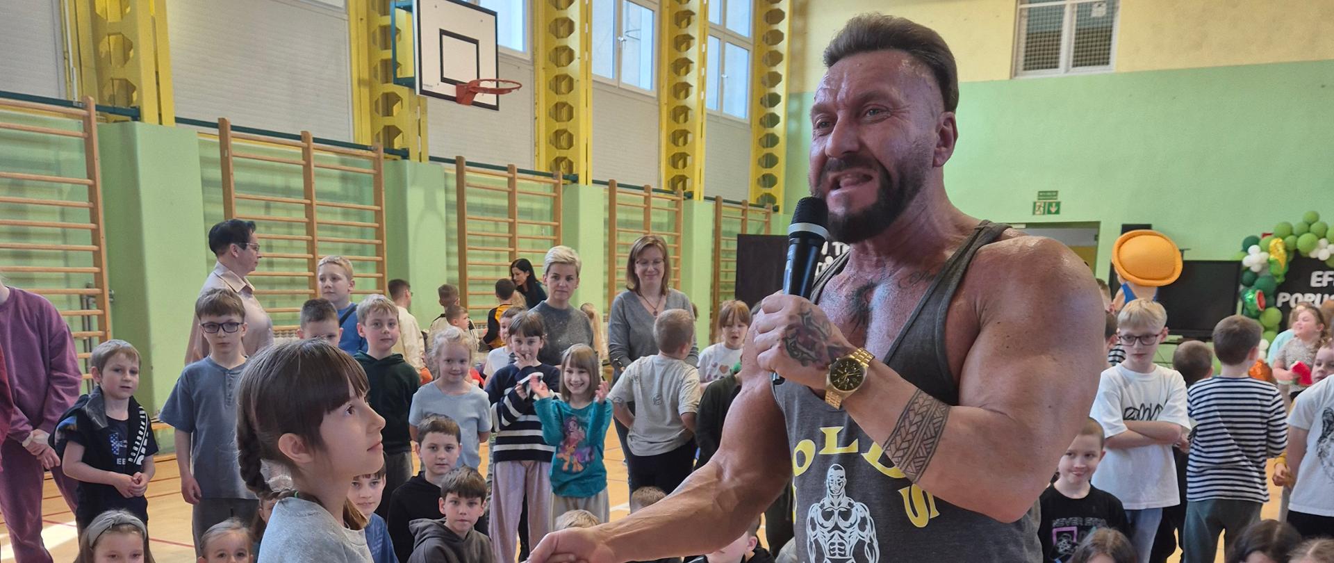 Mężczyzna w stroju sportowym, umięśniony. W prawym ręku trzyma mikrofon, lewą uściskuje dłoń dziewczynki stojącej przed nim. Za nimi duża ilość dzieci. Sala sportowa.