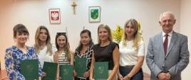 Grupowa fotografia w urzędzie przedstawiająca siedem osób stojących w rzędzie. Pięć kobiet po lewej stronie trzyma w dłoniach zielone teczki z pamiątkowym napisem i herbem Gminy Brańszczyk. Obok nich stoją kolejna kobieta oraz mężczyzna w szarej marynarce w kratę. Na białej ścianie w tle wiszą godło Polski, drewniany krzyż oraz herb gminy. W tle widoczne są drewniane biurka i bukiet słoneczników.