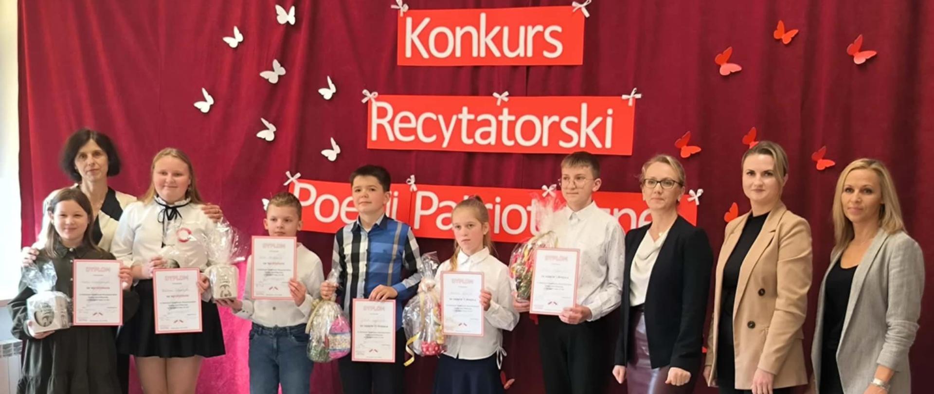 Nagrodzeni uczniowie, którzy wzięli udział w konkursie recytatorskim, ubrani na galowo, trzymają w ręku nagrody i dyplomy, w tle na bordowej kotarze biały napis "Konkurs Recytatorski Poezji Patriotycznej" oraz przypięte papierowe białe motylki