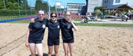 Trzy dziewczyny w czarnych bluzkach i czarnych spodenkach stoją na boisku do siatkówki plażowej na piasku. Stoją obok siebie i obejmują się w ramionach