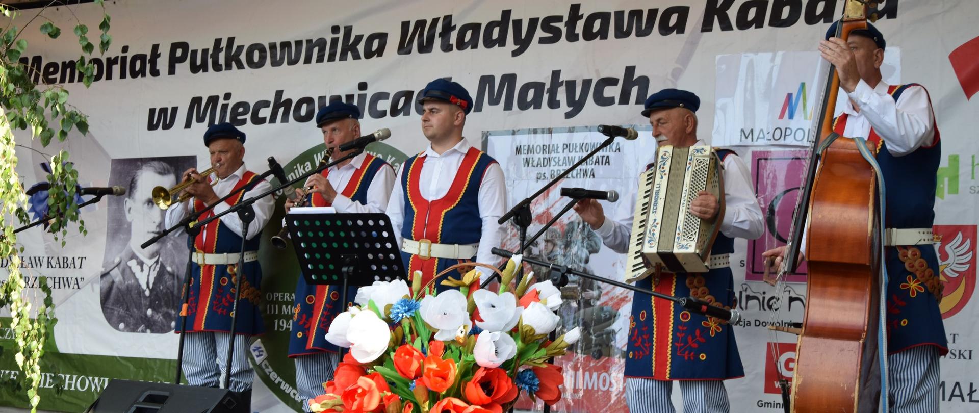 Na zdjęciu znajduje się pięciu mężczyzn w różnym wieku ubranych w tradycyjne stroje ludowe. Mężczyźni stoją na scenie i występują niektórzy trzymają instrumenty muzyczne takie jak: kontrabas, trąbka, akordeon i klarnet. W tle widnieje duży baner promocyjny wydarzenia. 