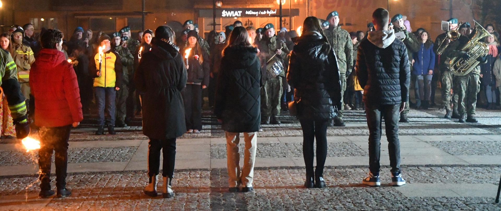 Wydarzenie na zewnątrz, publiczność wraz z wojskową orkiestrą stoją przed pomnikiem, przed nimi, zwróceni tyłem stoją dzieci trzymające pochodnie.