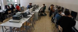 Zdjęcie przedstawia 13 osobową grupę uczniów korzystających z komputerów w pracowni komputerowej.