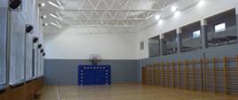 Duża sala sportowa z drewnianą posadzką i białym sufitem, wokół ścian barierki