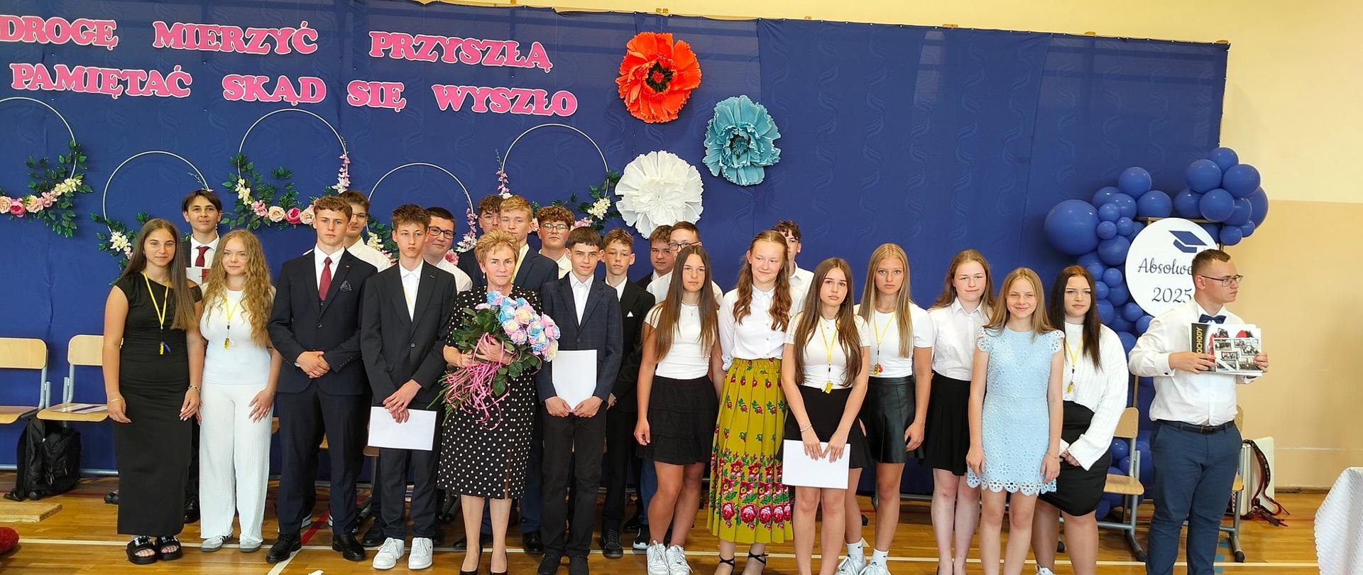 Cała klasa wraz z wychowawczynią, która otrzymała bukiet kwiatów, stoi na tle niebieskiej dekoracji z przypiętymi kwiatami i napisem "Aby w drogę mierzyć przyszłą, należy pamiętać, skąd się wyszło"