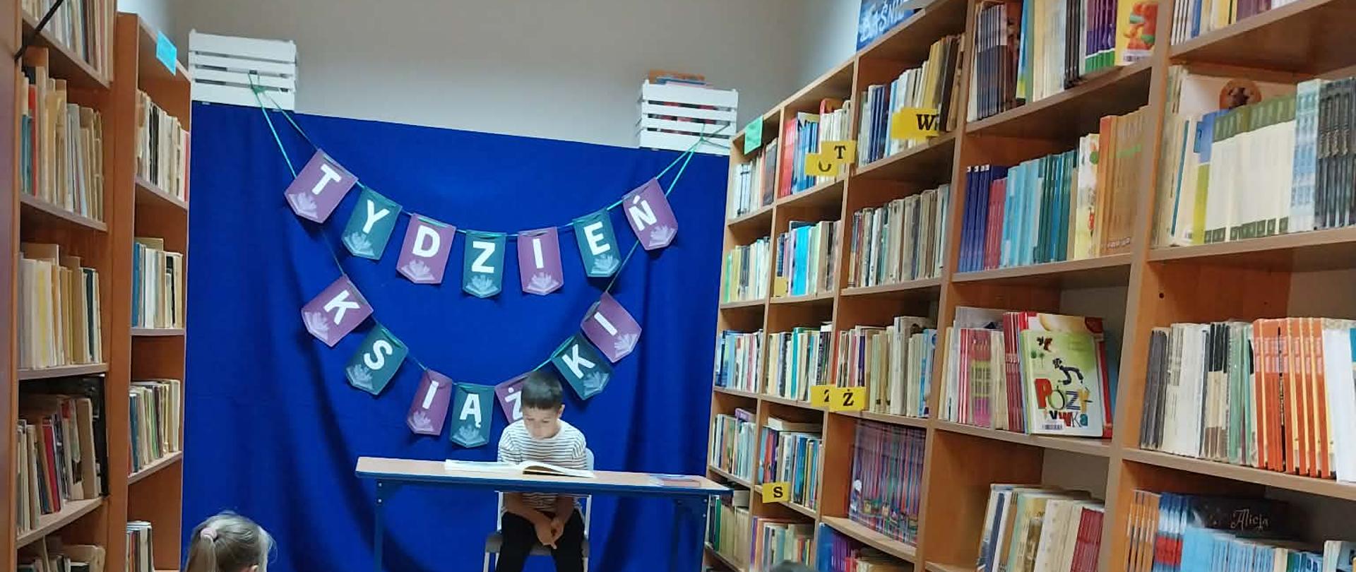 uczeń na tle napisu tydzień książki czyta książkę a przed nim siedzi widownia składająca się z jego kolegów i koleżanek
