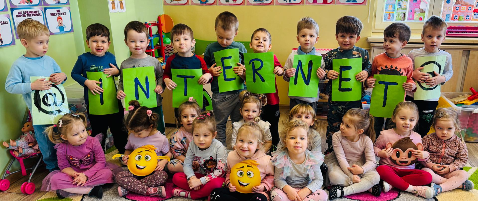 Grupa dzieci trzyletnich podczas Dnia Bezpiecznego Internetu stoi trzymając kartki z napisem „INTERNET”. Pozostałe dzieci siedzą na dywanie w pierwszym rzędzie, trzymając emotikony.