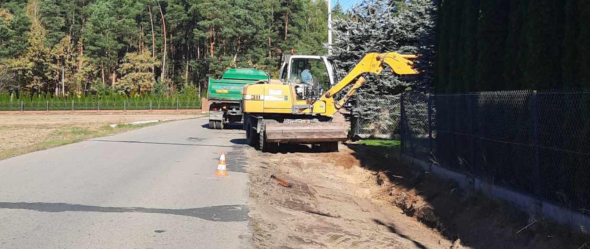 remont drogi asfaltowej, przekopane prawe pobocze w tle żółta koparka i zielony samochód ciężarowy 
