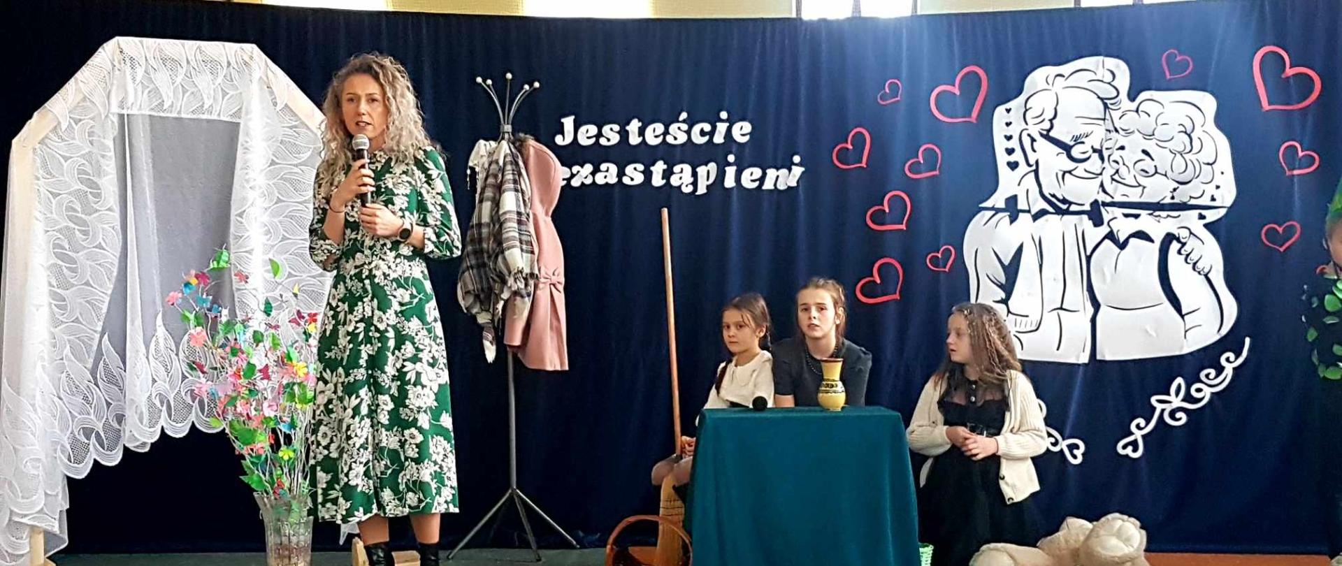 Tło przygotowane na uroczysty występ z okazji święta dziadków z cytatem "jesteście niezastąpieni". Na tle Pani Dyrektor w trakcie przemowy i powitania gości, obok przy stoliku trójka dzieci gotowych do inscenizacji.