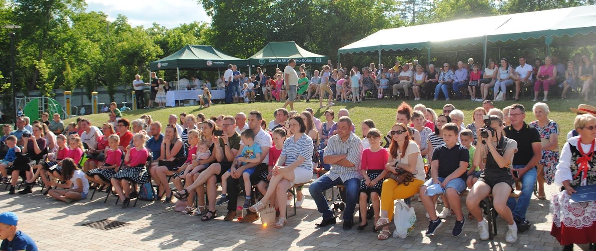 Zdjęcie przedstawia masę ludzi siedzących i stojących, oglądających występy artystyczne, pogoda jest słoneczna 