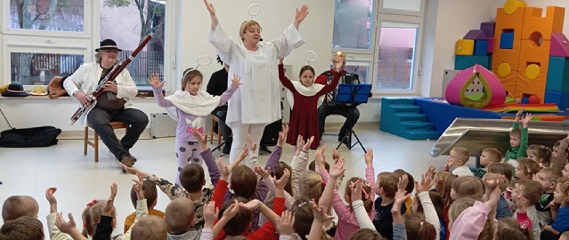 Zdjęcie prezentuje dzieci biorące udział w audycji muzycznej oraz teatrzyku. Przed grupą siedzących dzieci występują aktorzy
