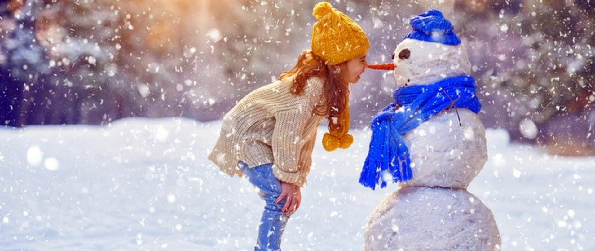 uśmiechnięta dziewczynka przystawia swój nos do nosa bałwana w aranżacji zimowej