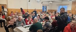 Grupa osób w różnym wieku siedzi przy stołach, oglądają występ grupy góralskiej.