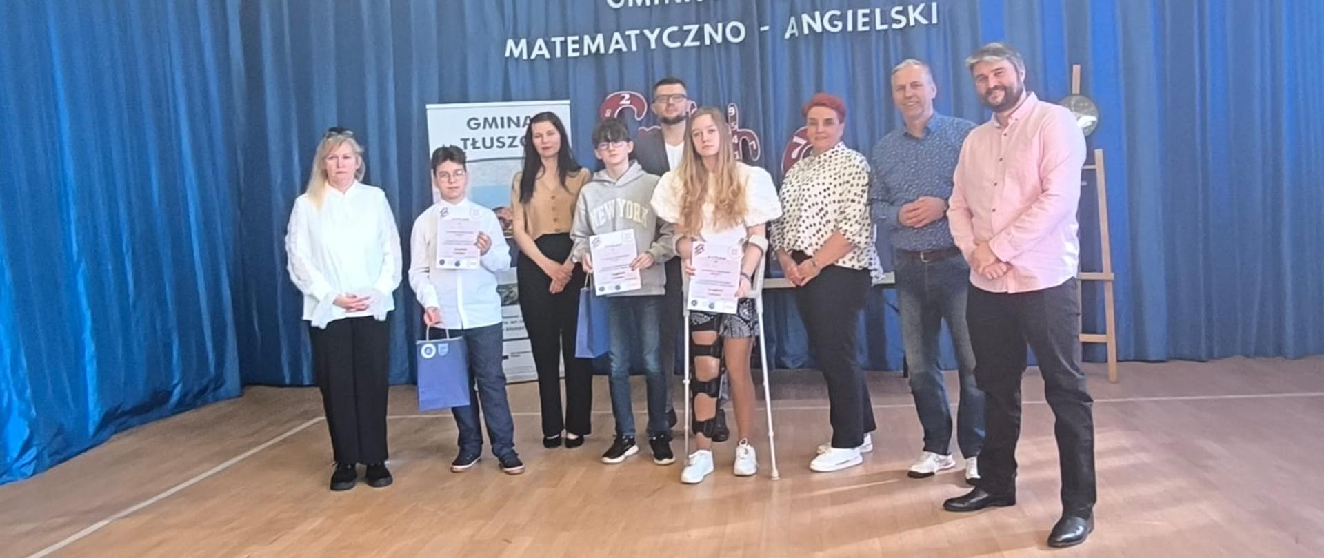 Grupa młodzieży, mężczyzn i kobieta, W środku stoi dziewczyna opierająca się na kulach z prawą nogą w ortezie. Za nimi napis Gminny Konkurs Matematyczno-Angielski 