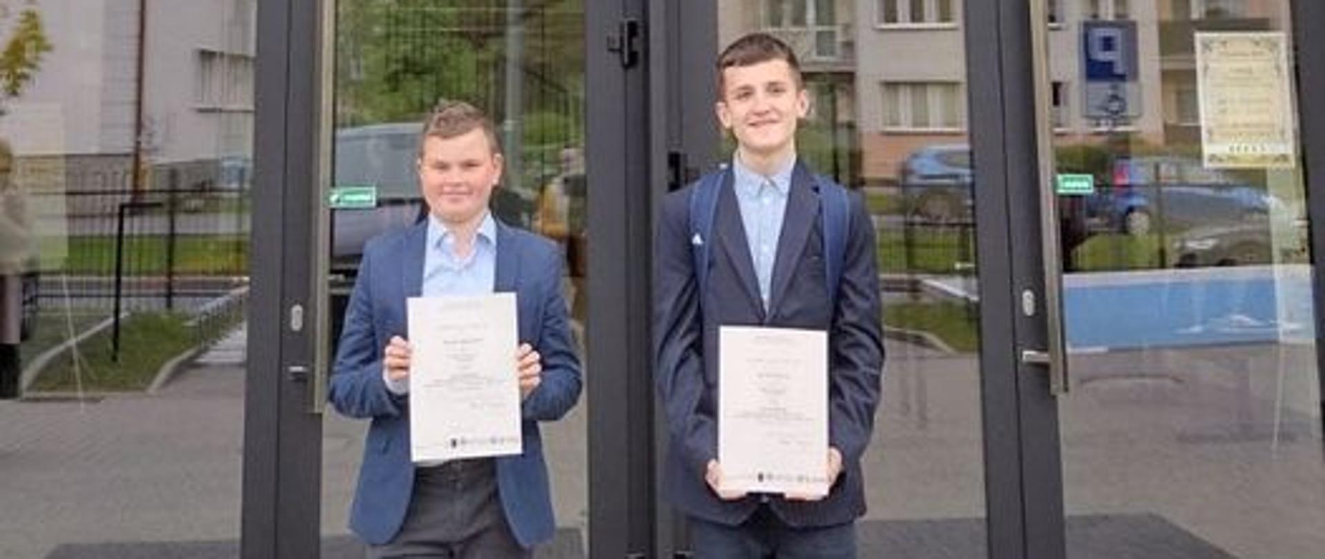 Przed wejściem głównym Akademii Nauk Stosowanych w Nowym Sączu stoją dwaj uczniowie ubrani odświętnie z dyplomami