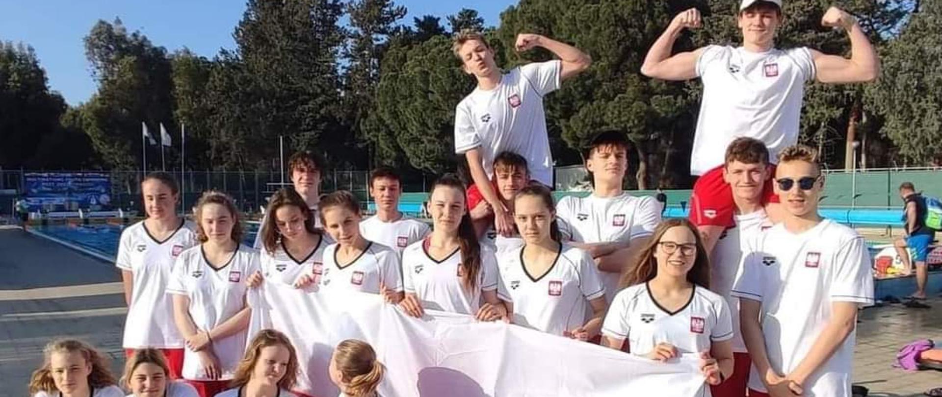 Zdjęcie przedstawia 19-osobową reprezentację Polski juniorów, stojącą na dworze przed odkrytą pływalnią. Zawodnicy pozują do zdjęcia ubrani w stroje sportowe w barwach biało - czerwonych. Pięciu pływaków trzyma w rękach rozłożoną flagę Polski