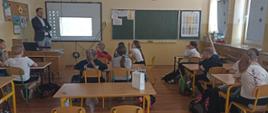 Koordynator programu "Aktywni Błękitni" stoi przy tablicy multimedialnej i przeprowadza prelekcję, uczniowie siedzą w ławkach i oglądają