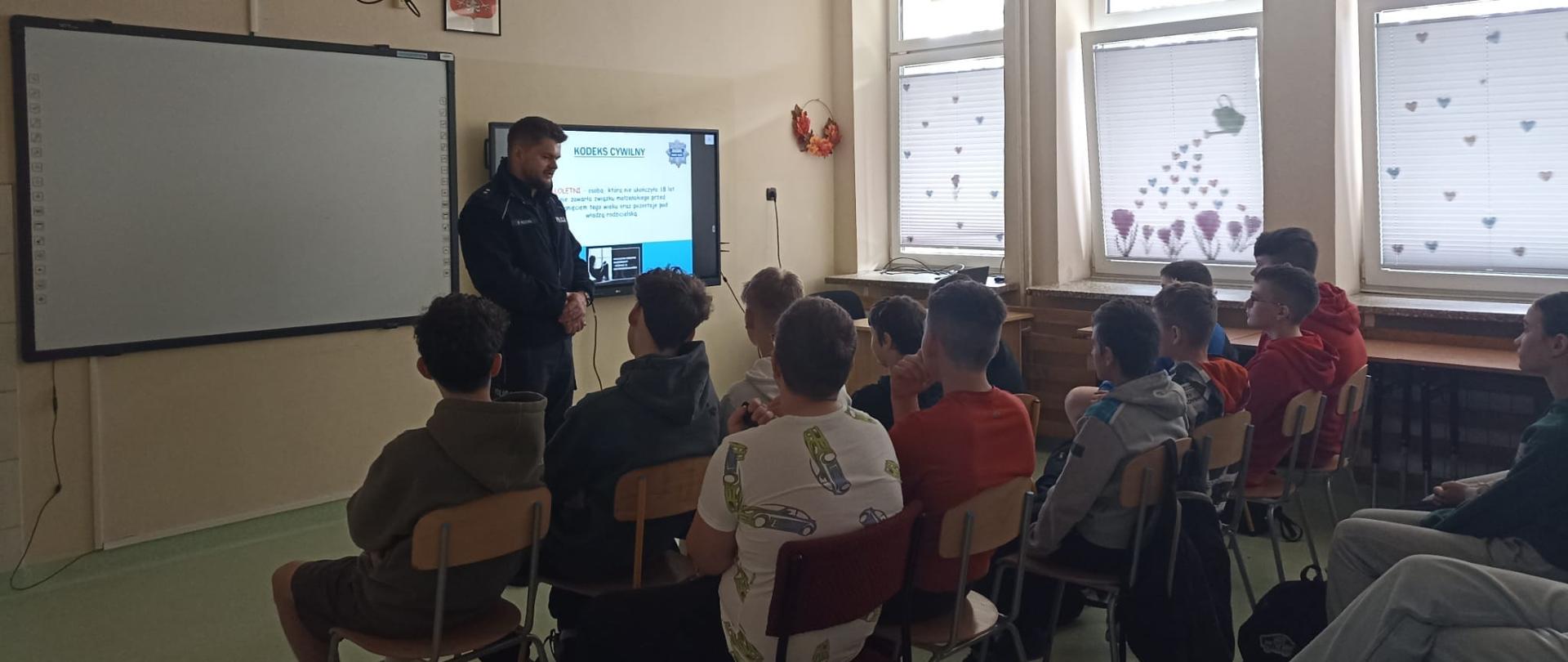 Uczniowie siedzą na krzesłach i słuchają prelekcji wygłaszanej przez policjanta, na tablicy interaktywnej wyświetlana jest prezentacja