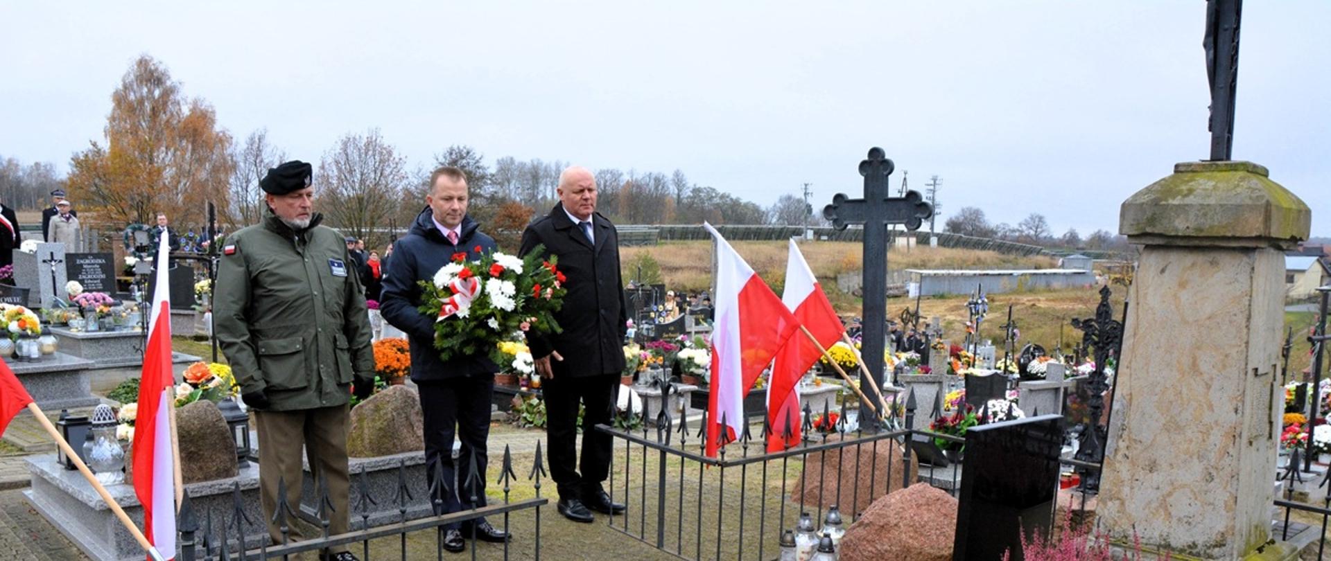 trzech mężczyzn w kurtkach stoją przed pomnikiem mężczyzna w środku trzyma wiązankę kwiatów biało czerwonych , 