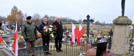 trzech mężczyzn w kurtkach stoją przed pomnikiem mężczyzna w środku trzyma wiązankę kwiatów biało czerwonych , 