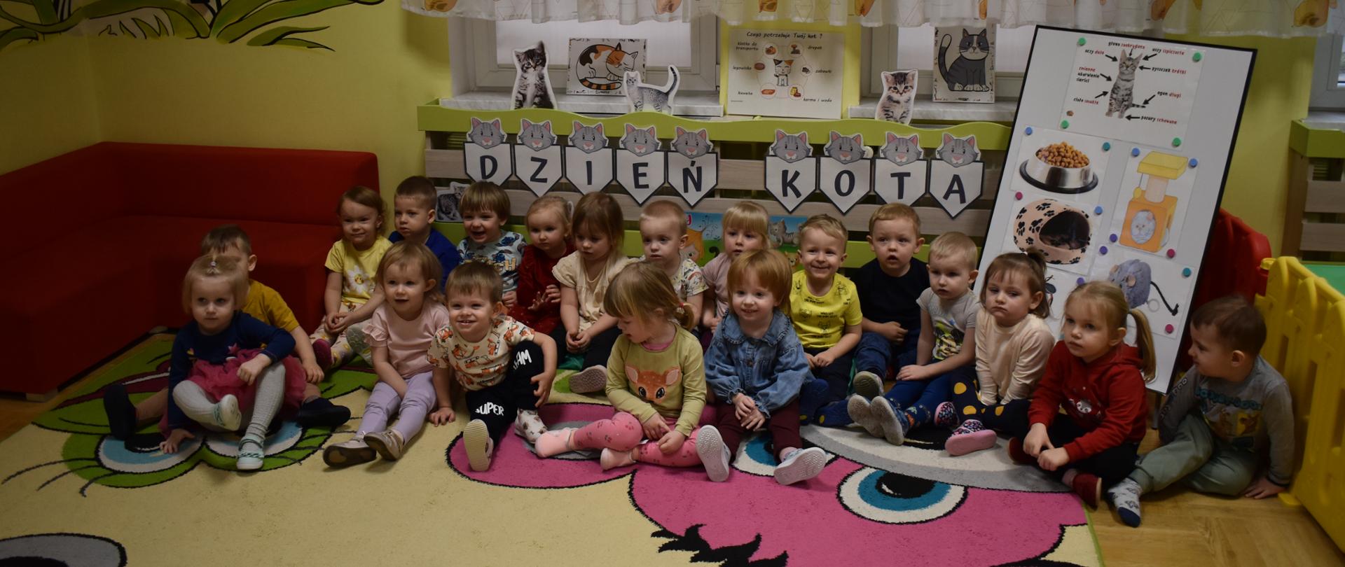 Na zdjęciu widać grupę dzieci siedzących na dywanie, w tle napis dzień kota i kocie akcesoria.