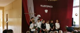 Uczniowie klasy czwartej biorą udział w uroczystej akademii z okazji rocznicy odzyskania niepodległości, na koszulach i bluzkach mają przypięte biało-czerwone kotyliony, w tle dekoracja patriotyczna 