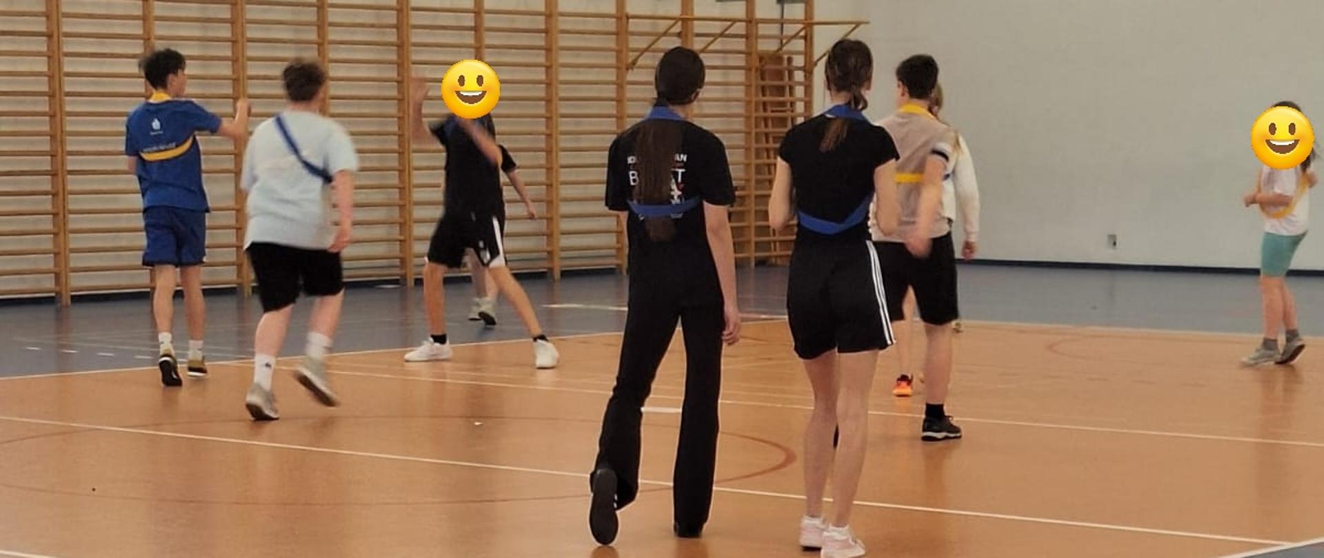 Na górze zdjęcia z lewej strony okna w białym kolorze, poniżej kosz zawieszony na ścianie oraz drabinki w kolorze jasnobrązowym. Na środku zdjęcia sylwetki osób ustawionych na boisku w różnych odległościach od siebie, ustawione tyłem. Na dole zdjęcia podłoga w kolorze niebieskim, z przedzielonymi czerwonymi liniami oraz białymi.