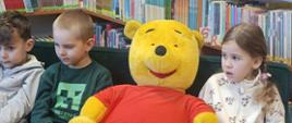 2 chłopców i dziewczynka siedzą na kanapie w bibliotece, między nimi duży żółty miś w czerwonym sweterku, z tyłu regały z książkami