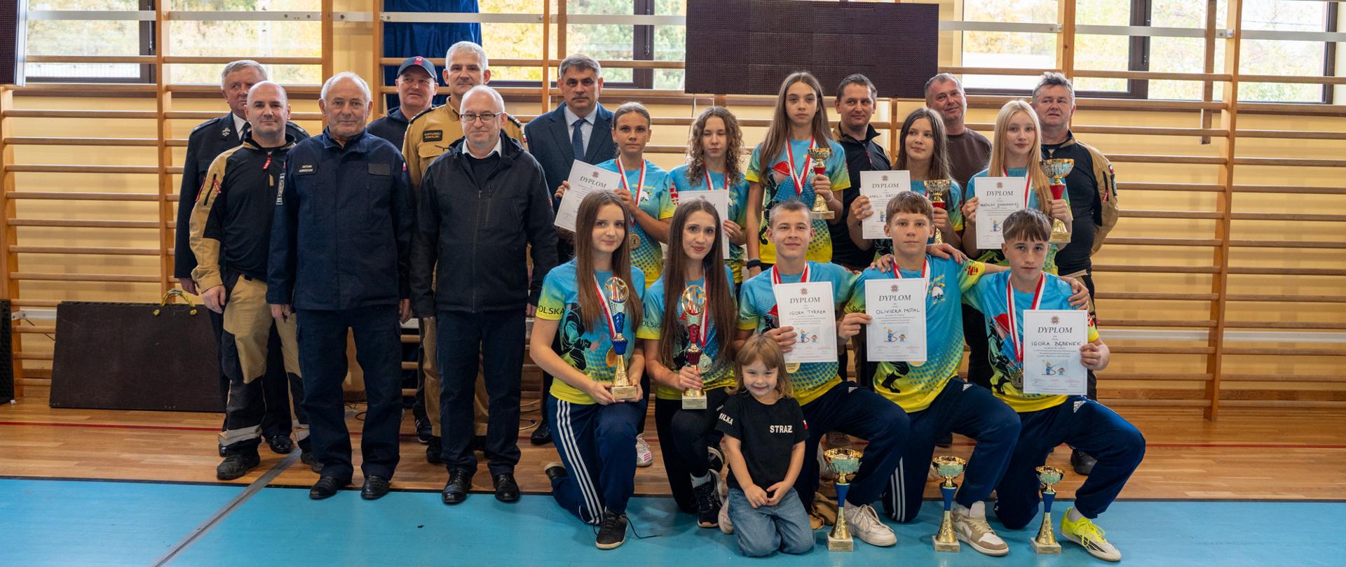 Zdjęcie grupowe uczestników i organizatorów Halowych Powiatowych Zawodów Młodzieżowych Drużyn Pożarniczych w Przytkowicach, które odbyły się 26 października 2026 roku. Na pierwszym planie młodzi zawodnicy w niebiesko-żółtych strojach sportowych prezentują zdobyte puchary, medale i dyplomy. Za nimi stoją przedstawiciele straży pożarnej, organizatorzy oraz zaproszeni goście. Fotografia została wykonana w hali sportowej, na tle drabinek gimnastycznych i baneru zawodów.