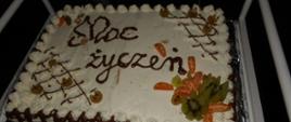 Prostokątny tort pokryty białą bitą śmietaną i dekoracjami z ciemnej czekolady oraz świeżymi owocami. Na torcie widoczny jest napis: "Moc życzeń".