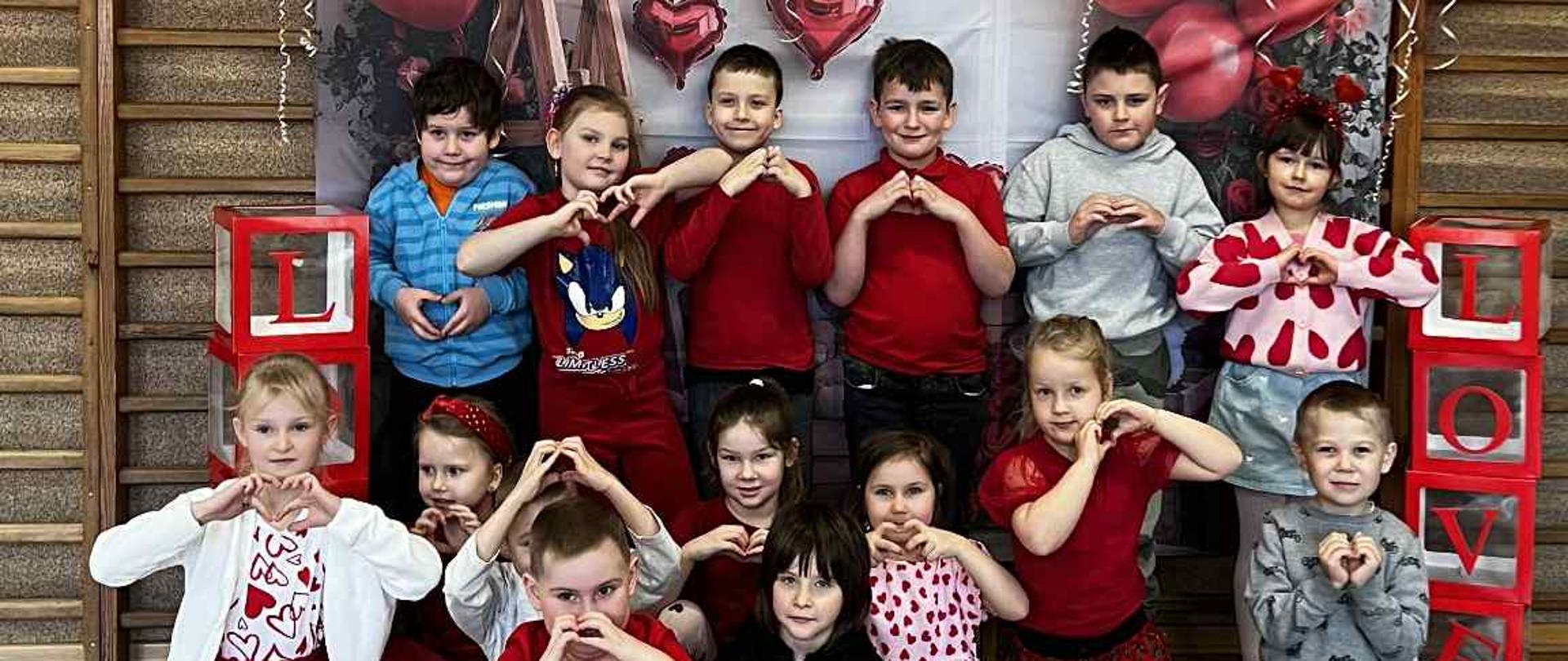 Dzieci z Oddziału Przedszkolnego A pozują do wspólnego zdjęcia na specjalnie przygotowanej ściance walentynkowej. Za nimi, na drabinkach wiszą czerwone balony w kształcie serc oraz kartoniki z literami, tworzącymi napis: LOVE.
