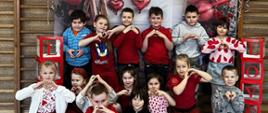 Dzieci z Oddziału Przedszkolnego A pozują do wspólnego zdjęcia na specjalnie przygotowanej ściance walentynkowej. Za nimi, na drabinkach wiszą czerwone balony w kształcie serc oraz kartoniki z literami, tworzącymi napis: LOVE.