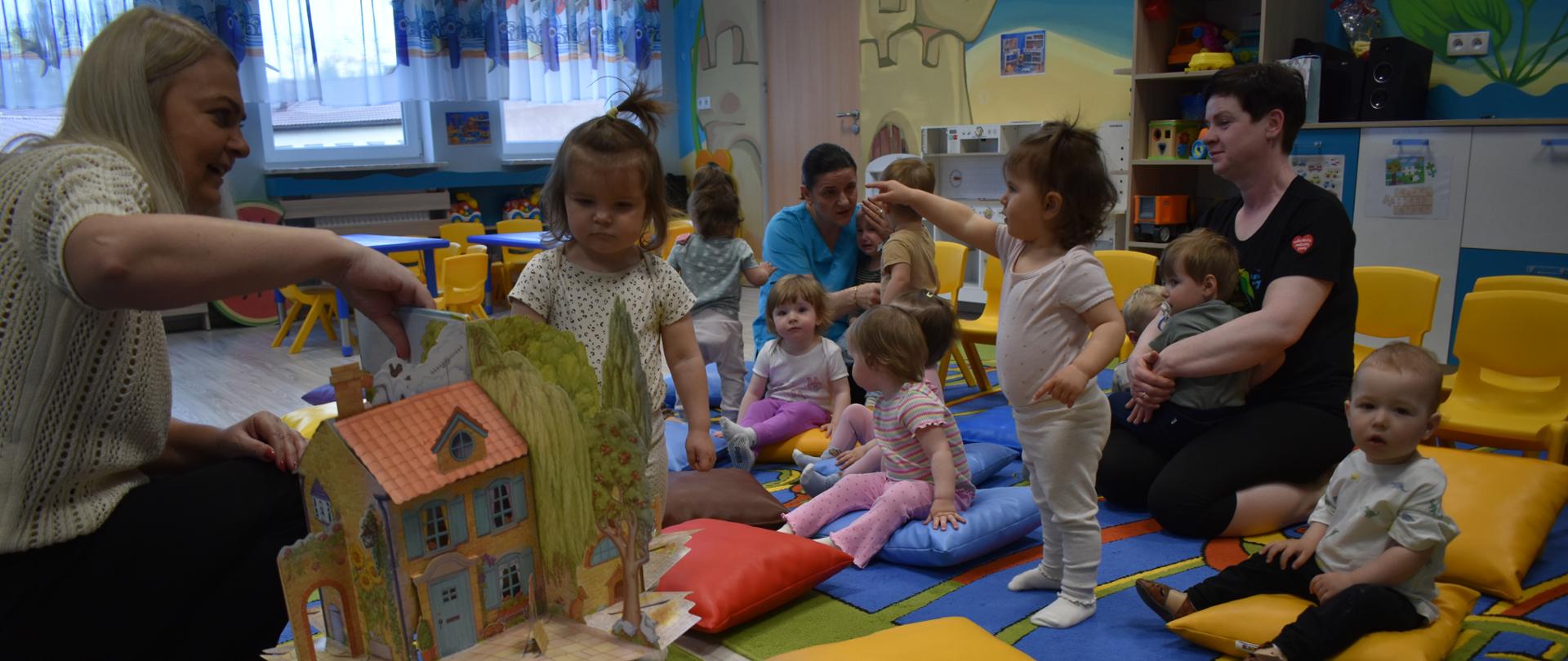 Na zdjęciu są dzieci z grupy Słoneczka oraz pani z Biblioteki Pedagogicznej im. Heleny Radlińskiej w Sokołowie Podlaskim. Dzieci oglądają pokaz książek przygotowanych przez zaproszonego gościa.
