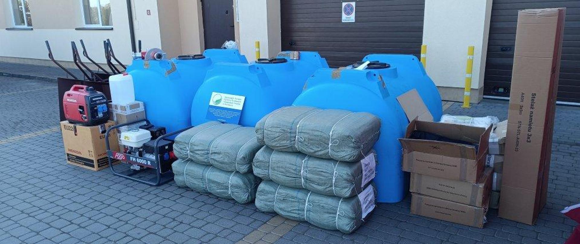 5 pakunków owiniętych sznurkiem, 34 duże niebieskie zbiorniki, po prawej kartony, po lewej agregat i brązowe taczki
