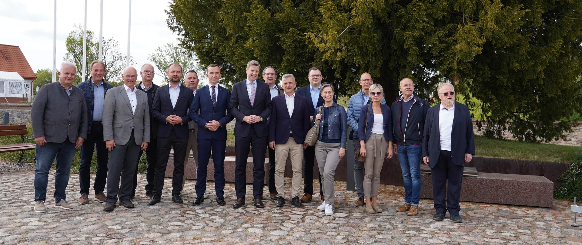 Grupa ludzi stojąca na brukowanym dziedzińcu. W tle po lewej stronie flagi Polski, UE i miasta Lubawy, a po prawej drzewa. 