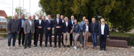 Grupa ludzi stojąca na brukowanym dziedzińcu. W tle po lewej stronie flagi Polski, UE i miasta Lubawy, a po prawej drzewa. 