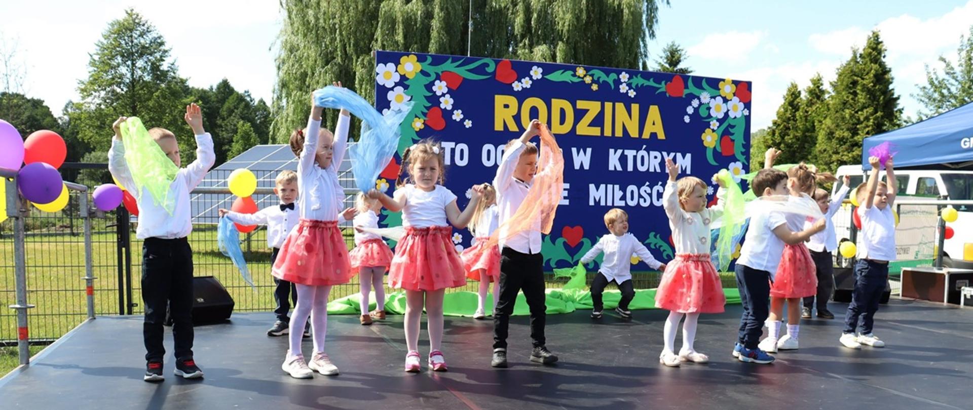 dziewczynki w białych bluzeczkach i różowych spódniczkach a chłopcy w białych koszulach i czarnych spodniach wspólnie tańczą na scenie 