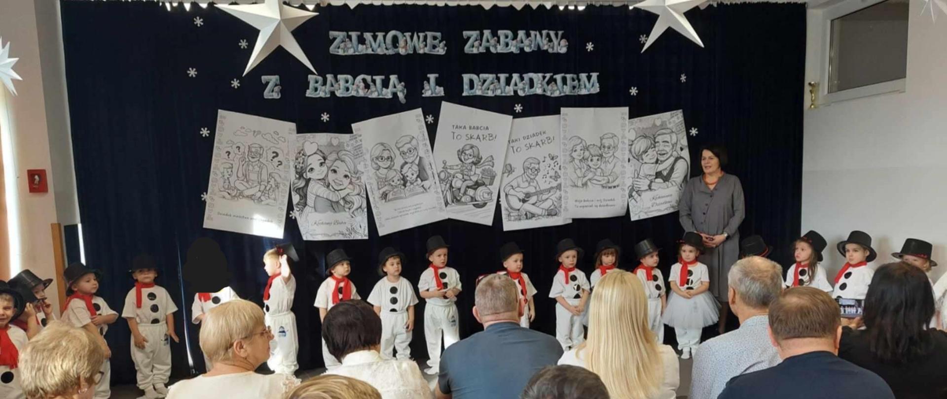 Pani Dyrektor stoi pomiędzy dziećmi przebranymi za bałwanki przemawiając do zebranych gości przed rozpoczęciem uroczystości. Na zdjęciu prezentuje się cała grupa Fajniaków. 