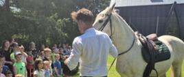 Na trawce przy szkole właściciel prezentuje zgromadzonym dzieciakom białego konia