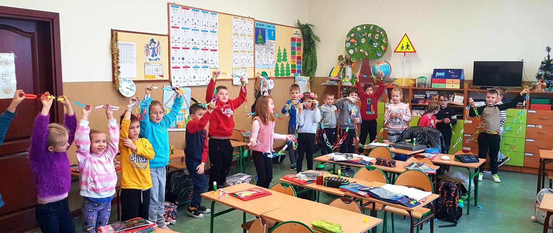 Uczniowie najmłodszej klasy naszej szkoły, ustawieni w półkole trzymają wysoko nad głową łańcuch zbudowany z kolorowych kawałków papieru. Na twarzach uczniów widnieje uśmiech i zadowolenie.
