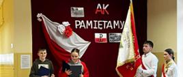 Uczennica i uczeń odczytują teksty, po prawej stronie stoi poczet sztandarowy szkoły ze sztandarem, za nimi bordowa zasłona, na której przypięto biało czerwoną flagę, napis "AK pamiętamy" oraz symbol Polski walczącej, na pierwszym planie widać uczniów oglądających akademię