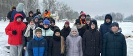 Grupa uczniów pozuje do wspólnego zdjęcia. Wszyscy ubrani są w ciepłe, zimowe kurtki i stoją na ośnieżonym chodniku. Za nimi znajduje się zamarznięta rzeka oraz ratownicy WOPR.
