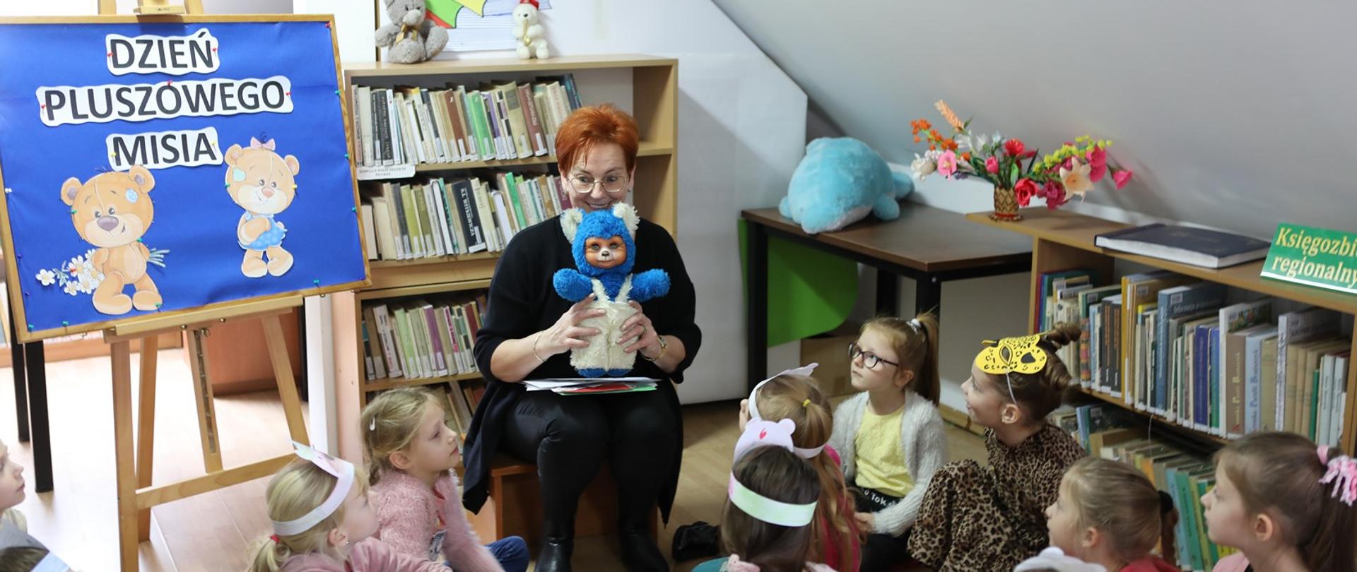 Pani bibliotekarka trzymająca w rękach maskotkę, przed nią na podłodze siedzi gromadka dzieci.