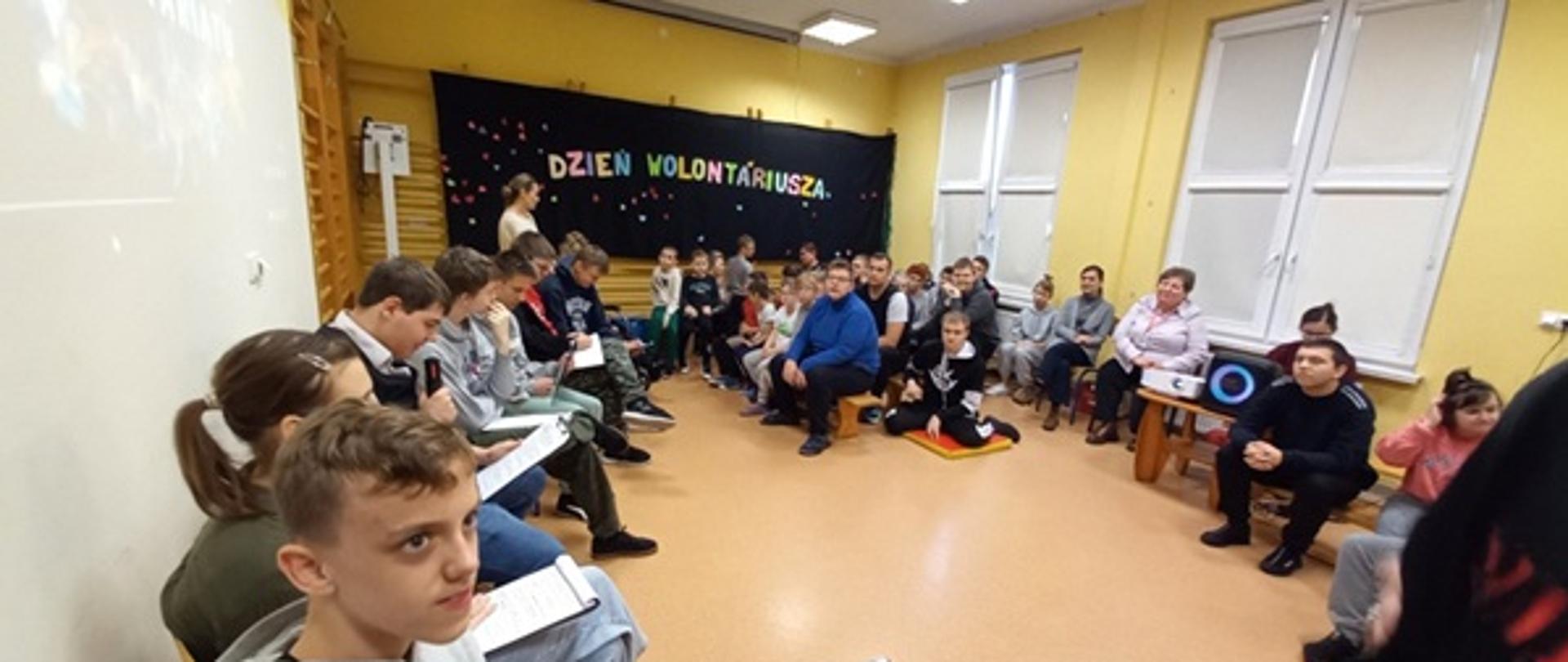 Uczniowie siedzą w półkolu na Sali gimnastycznej i rozmawiają o wolontariacie i wolontariuszach. Na ścianie widoczny jest kolorowy napis „ Dzień Wolontariusza”