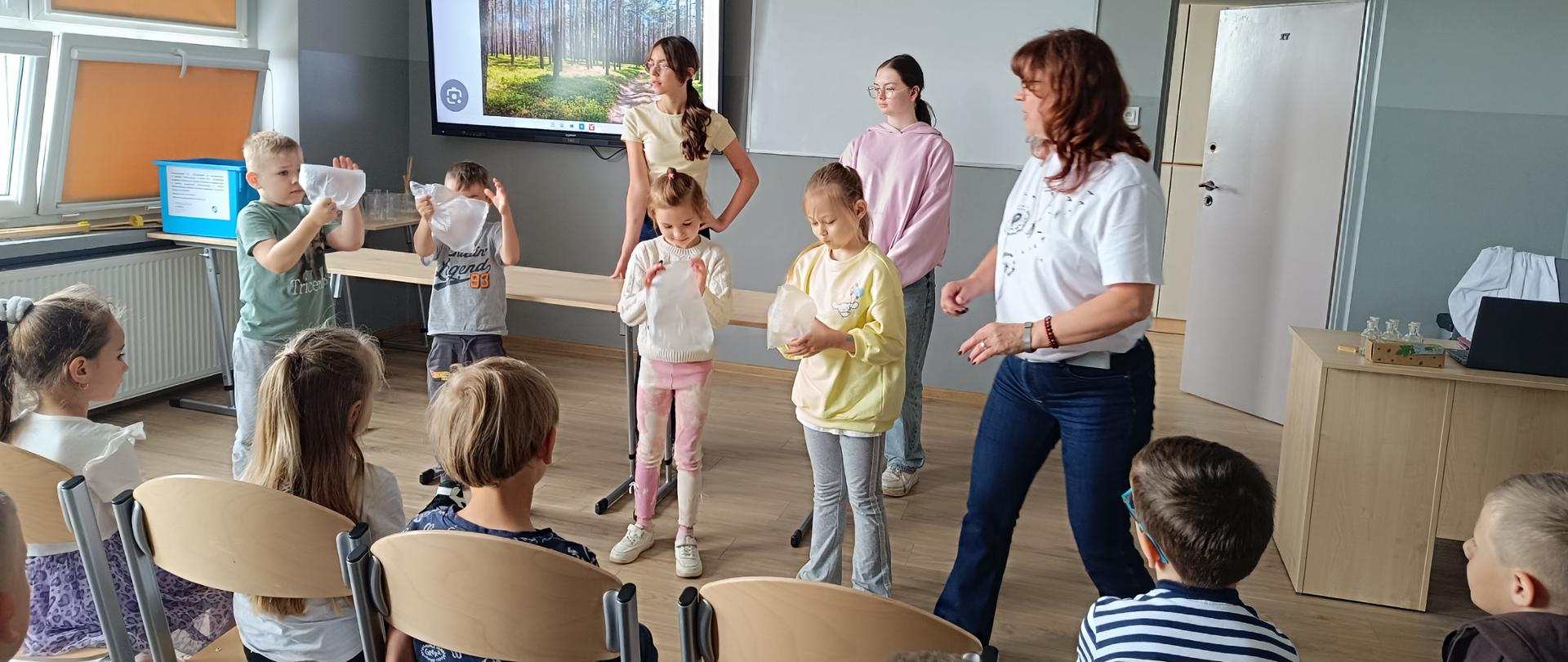 Duża sala w której na krzesłach siedzą dzieci w wieku przedszkolnym odwrócone tyłem do oglądającego zdjęcie. Przed nimi stoi czworo dzieci, trzymają woreczki napompowane powietrzem, po prawej stronie kobieta w białej bluzce i jeansach. Za nimi stoją stoliki i biurko oraz dwie młode dziewczyny. Po lewej stronie duże okno zasłonięte brązową roletą. Na wprost duży ekran monitora z krajobrazem lasu, biała tablica oraz drzwi. 