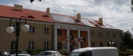 Budynek Urzędu Gminy w Pokrzywnicy z czerwonym dachem i beżową elewacją. Na dachu zamontowane są panele fotowoltaiczne. Przed budynkiem zaparkowane są samochody osobowe i biały van. W kadrze widoczne są latarnie z kulistymi kloszami oraz gałęzie drzew w górnej części zdjęcia. Na tle nieba widać chmury z przejaśnieniami.