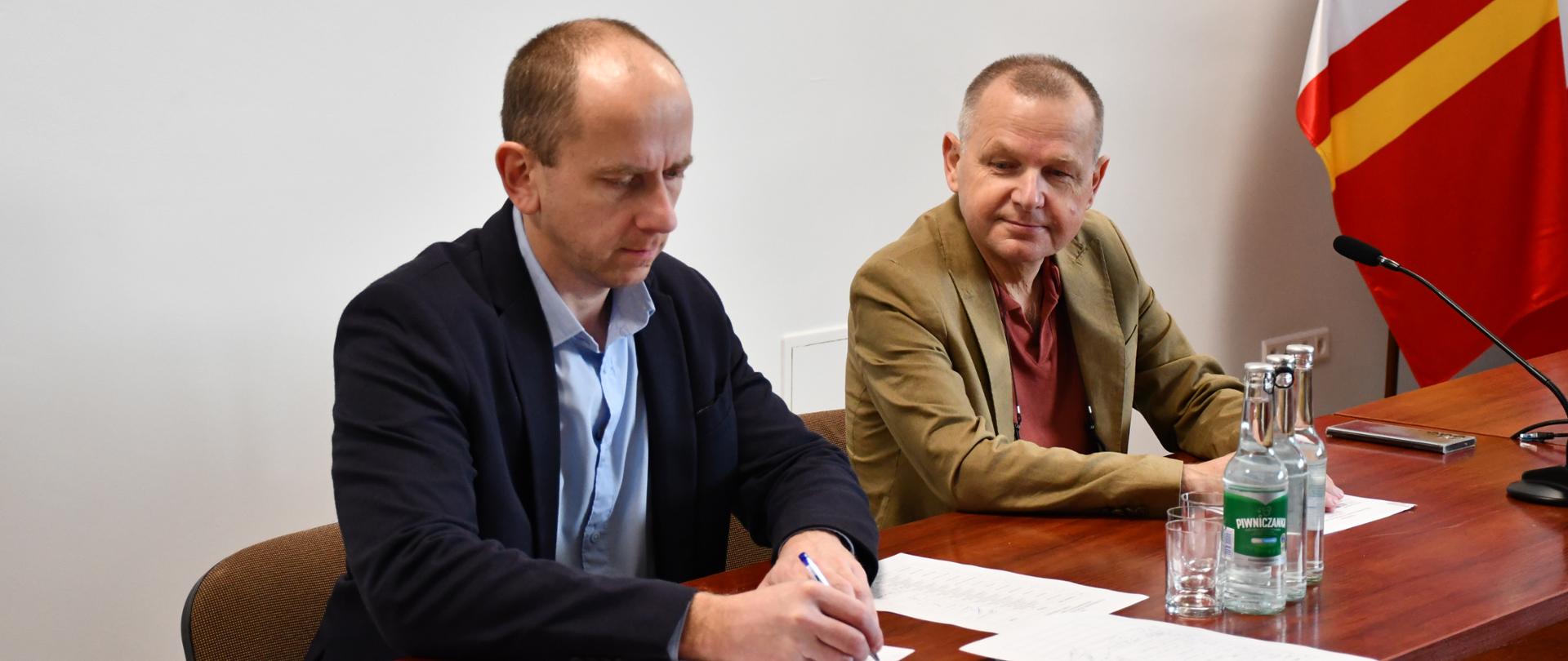Przy stole obok siebie siedzą Starosta Dąbrowski i pracownik Referatu Zarządzania Kryzysowego i Spraw Obronnych